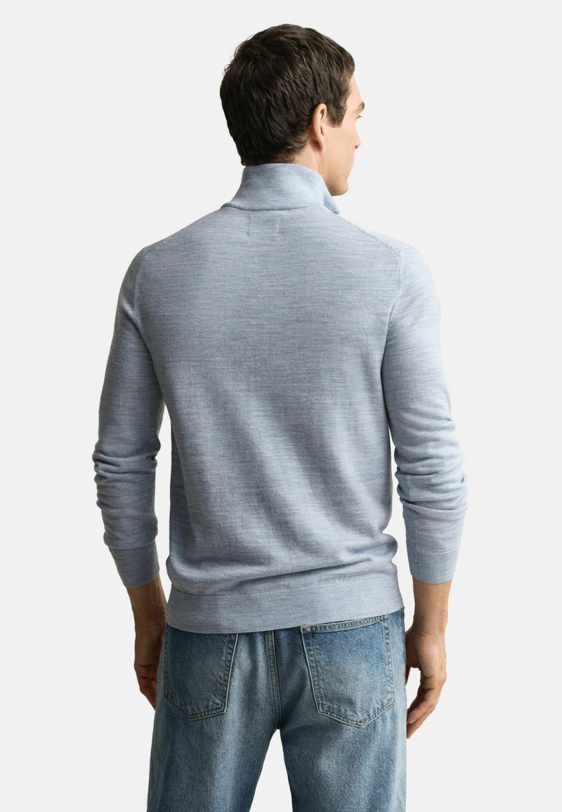 Ein Mann trägt einen hellblauen Rollkragensweater und blaue Jeans, von hinten betrachtet vor einem schlichten weißen Hintergrund.