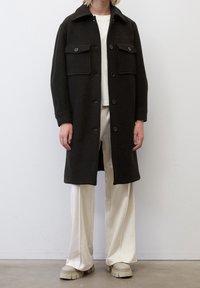 Cappotto di lana scuro con colletto, chiusura a bottoni e due tasche frontali; indossato sopra una camicia chiara e pantaloni color crema, con scarpe chunky beige.