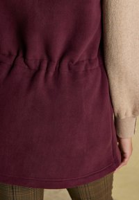 Bordeauxfarbene Fleece-Oberbekleidung mit geschnürtem Taillendetail, kombiniert mit einem beigen strukturierten Ärmel; am Boden ist der gemusterte Hosenumen sichtbar.