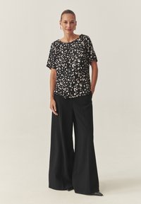 Fekete rövid ujjú felső világos bézs absztrakt mintával, magas derekú fekete bő szárú nadrággal és fekete hegyes orrú cipőkkel párosítva.