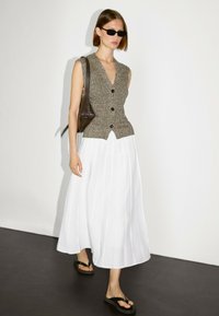 Frau trägt einen ärmellosen Strickpullunder, einen weißen, plissierten Midirock, schwarze Flip-Flops, Sonnenbrille und hält eine kleine dunkle Schultertasche.