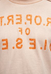 Μπεζ ύφασμα με ξεθωριασμένο πορτοκαλί κείμενο τυπωμένο ανάποδα, με μερικά γράμματα και λέξεις να επαναλαμβάνονται σε κυρτή διάταξη.