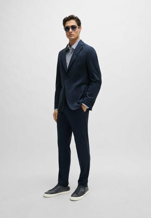 Completo blu con una vestibilità su misura, abbinato a una camicia chiara a fantasia. Accentuato da sneakers scure e occhiali da sole. Tessuto morbido.