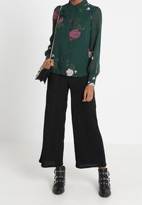 Personne portant une blouse florale vert foncé, un pantalon large noir, des bottines noires à clous, tenant un petit sac noir avec une bandoulière en chaîne.