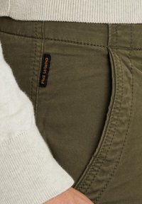 Olivgrüne Stretchhose mit einer Seitentasche, aus strukturiertem Stoff und einem schwarzen Etikett mit "Pure Legend" in orangefarbener Ziernaht.