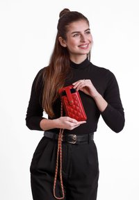 Červená prošívaná crossbody peněženka na telefon s zlatým zapínáním a řetízkovým popruhem. Má elegantní obdélníkový tvar a hladkou texturu.