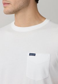 Camiseta blanca de algodón con cuello redondo, que presenta un bolsillo en el pecho con una etiqueta azul marino. Textura suave y diseño sencillo.