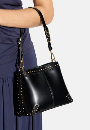 Osoba ubrana w ciemnofioletowy strój trzymająca czarną skórzaną torebkę z złotymi ćwiekami i regulowanym paskiem na przedramieniu.