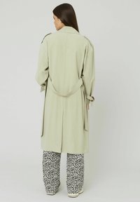 Manteau de trench vert clair avec une taille ceinturée, arborant une coupe décontractée et des poches latérales, associé à un pantalon à larges jambes à pois noirs et blancs.