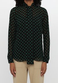 Femme portant un chemisier noir transparent à pois verts et un nœud papillon noué au cou, associé à un pantalon beige avec des poches plaquées devant.