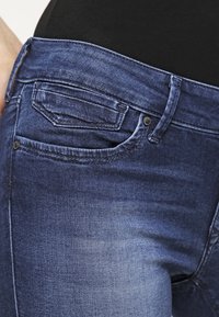 Mörkblå jeans med en slät textur, med en bakficka, nitar och en standard midjeband. Ingen synlig slitning eller mönster.