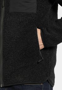 Mustavillainen fleecetakki, jossa on teksturoitu pinta, vetoketjullinen sivutasku ja sileä kangasosio rinnassa, viimeistelty kontrastivärisin sauman.