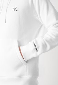 Sudadera blanca con capucha que presenta un pequeño logotipo negro "cK" en el pecho y el texto "CALVIN KLEIN JEANS" en el puño. Textura suave, corte regular.