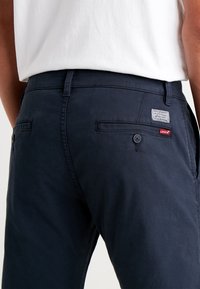 Tmavě modré bavlněné kalhoty s zadními kapsami, zapínáním na knoflíky a štítkem s logem "Levi's". Materiál má hladkou texturu.