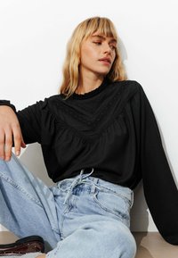 Haut noir à manches longues en tissu texturé avec un col froncé et des coutures décoratives, associé à un jean bleu clair.