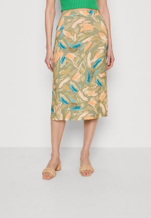 Mujer con una falda midi con estampado abstracto en beige, azul y durazno, sandalias tejidas beige con tacón y una camiseta verde.