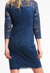 Robe en dentelle bleu marine avec des manches trois-quarts, des motifs floraux et un ourlet en forme de vagues. Le tissu présente un design texturé et complexe.