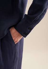 Chemise en lin bleu marine avec un gros plan d'une main dans une poche. Présente un poignet à boutons avec deux boutons foncés sur une manche structurée.