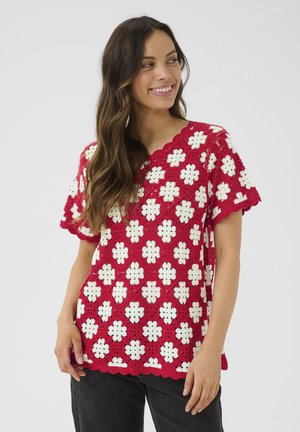 Haut en crochet rouge avec des motifs floraux blancs, des bords festonnés et des manches courtes, associé à un pantalon foncé. Design ouvert et texturé.