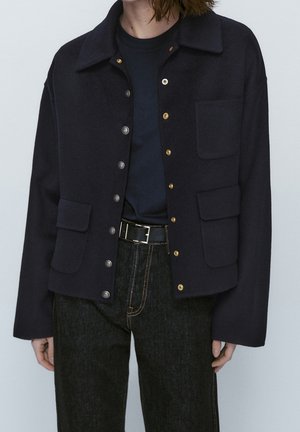 Person wearing a black cropped jacket with mixed metal buttons, patch pockets, black belt, dark jeans, and a black t-shirt beneath.