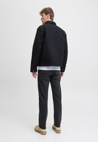 Chaqueta negra con cuello, usada sobre una camisa blanca. Pantalones gris oscuro y zapatos beige completan el conjunto. Fondo liso.