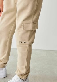 Beige kokvilnas bikses ar sānu kabatu un ar druku "BAKER by Ted Baker" tuvu gurnam, apvienotas ar baltām sporta kedām.