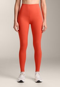 Magas derekú, testhezálló leggings élénk piros színben. Rugalmas anyagból készül, sima tapintással, fehér sportcipőkkel párosítva.