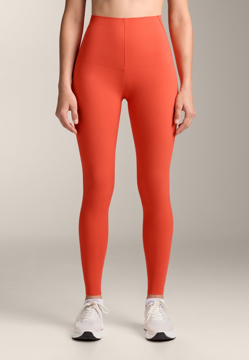 Magas derekú, testhezálló leggings élénk piros színben. Rugalmas anyagból készül, sima tapintással, fehér sportcipőkkel párosítva.