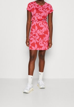 Femme portant une robe rouge à manches courtes avec de grands motifs floraux roses, des chaussettes blanches et des baskets blanches, debout devant un fond blanc.