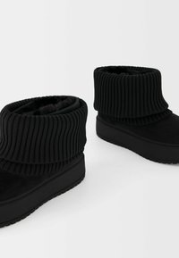 Bottes noires aux chevilles avec une manche en tricot côtelé et doublure en fausse fourrure, présentant une semelle plate-forme noire unie et une texture extérieure lisse.