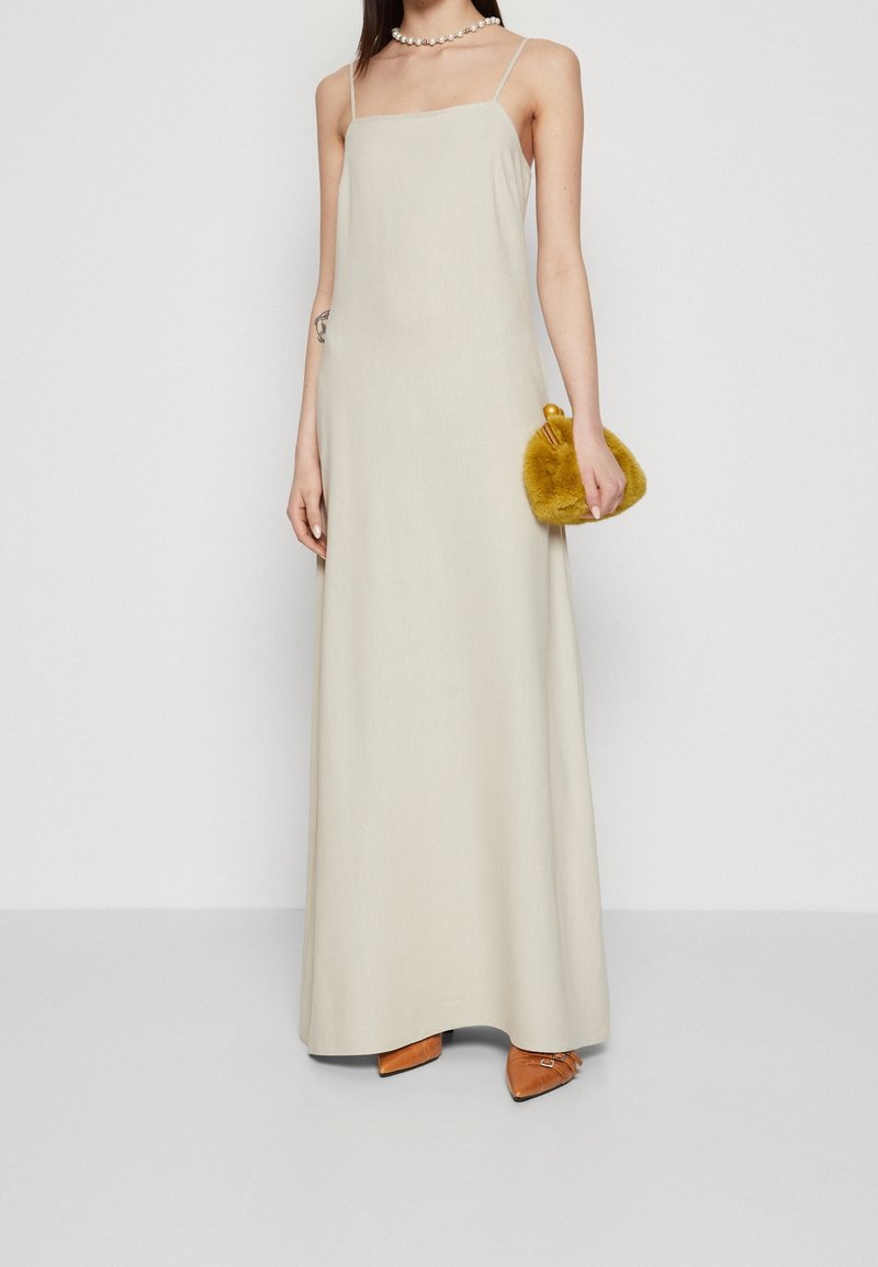 Femme portant une longue robe beige sans manches, tenant un petit sac à main pelucheux jaune moutarde et portant des chaussures marron pointues.