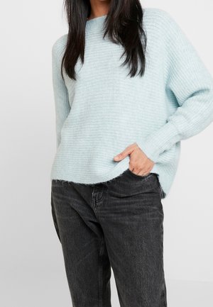 Maglione a maglia testurizzata di colore azzurro chiaro con scollo rotondo e spalle abbassate, abbinato a jeans grigi scuri. Il maglione presenta una parte posteriore leggermente più lunga.