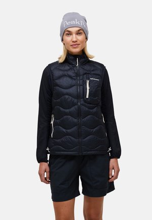 Kvinde iført en grå hue, sort quiltet vest med lynlåslommer, sort bluse med lange ærmer og sorte shorts mod en ensfarvet baggrund.
