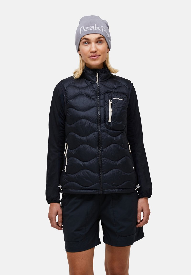Femme portant un bonnet gris, un gilet matelassé noir avec poches zippées, un haut noir à manches longues et un short noir sur un fond uni.