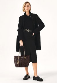 Bruine tote bag met krokodillendessin, zilveren kettinghandvaten, sleutelhanger met franjes en geëtst logo. Model draagt een geheel zwarte outfit en een jas.