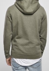 Olivgrön huvtröja med mjuk textur, justerbar huva och ribbade muddar. Bärs över en vit skjorta och ihop med ljusblå jeans.