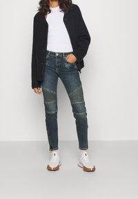 Tmavě modré džíny s žebrovanými detaily na kolenou, se zipovými uzávěry a slim fit, doplněné černým kardiganem a bílými sportovními teniskami.