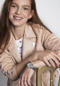 Lichtroze suède jasje, ronde zilveren watch met een stenen bezel, gelaagde armbanden en ringen. Lang, stijl haar, neutrale makeup.