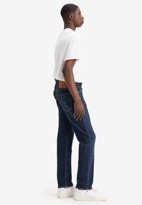 Jeans de mezclilla azul oscuro con diseño de pierna recta, que cuentan con un parche de cuero marrón en la parte posterior, combinados con una camiseta blanca de manga corta sencilla.