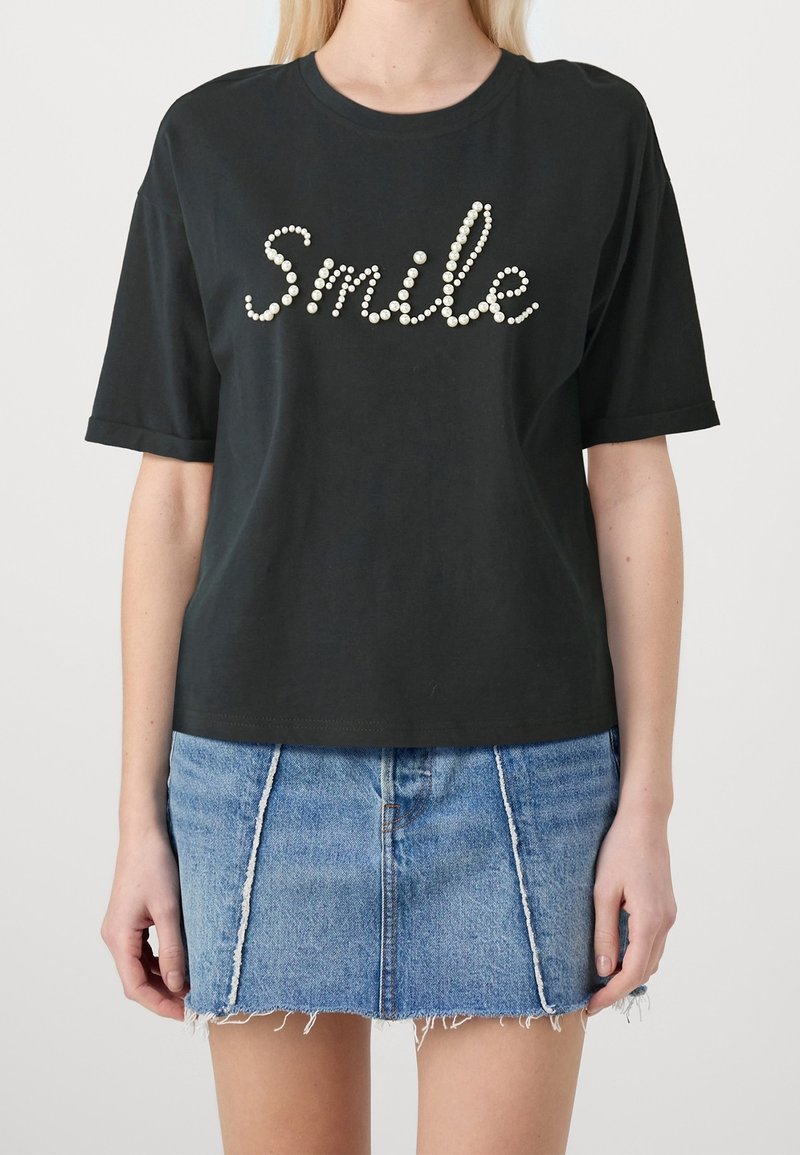 Sort bomulds-T-shirt med korte ærmer, der har et "Smile"-tekstdesign lavet af hvide perle-lignende pynt. Bæres med et denimskørt.