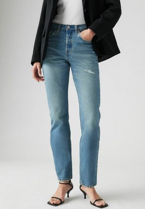 Hellblaue High-Waist-Jeans mit lockerem Schnitt, die eine ausgewaschene Optik und ein kleines, destriertes Detail an der Vorderseite aufweisen, kombiniert mit schwarzen Absätzen.