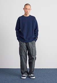 Marineblauwe sweatshirt met geribde manchetten, gecombineerd met olijfgroen cargobroek met meerdere zakken. Draagt zwarte en witte sneakers.