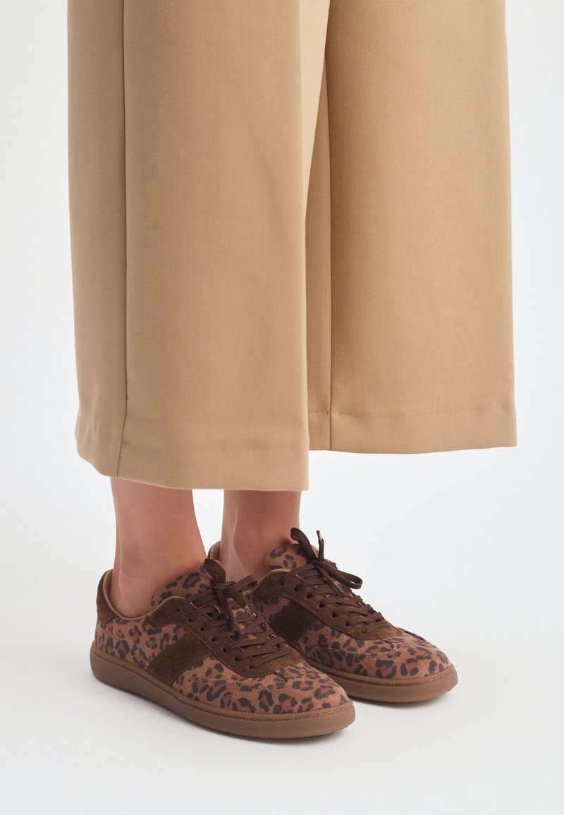 Brązowe zamszowe sneakersy z leopardzim wzorem, teksturowymi akcentami i brązowymi sznurówkami, w zestawieniu z beżowymi spodniami typu wide-leg.