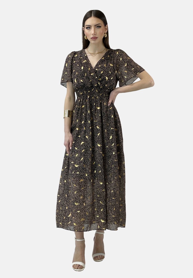 Robe maxi en chiffon noir avec un motif de feuilles dorées, manches courtes, décolleté en V, taille élastique et jupe fluide, assortie à des talons blancs à lanières.