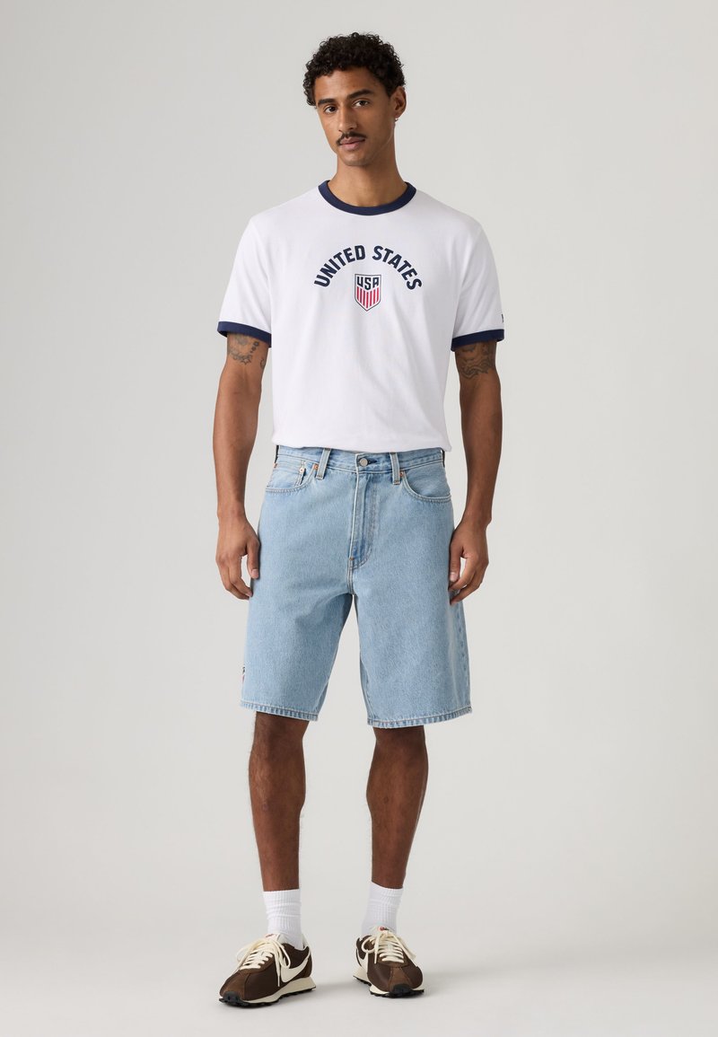 Mann trägt ein weißes „United States“ T-Shirt mit marineblauen Rändern, hellblaue Jeansshorts, weiße Socken und braune Sneakers und steht vor hellem Hintergrund.