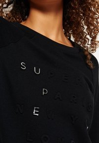 Persona dai capelli ricci che indossa un felpa nera con lettere bianche e nere in rilievo che scrivono: Super, Parigi, New York, Londra.