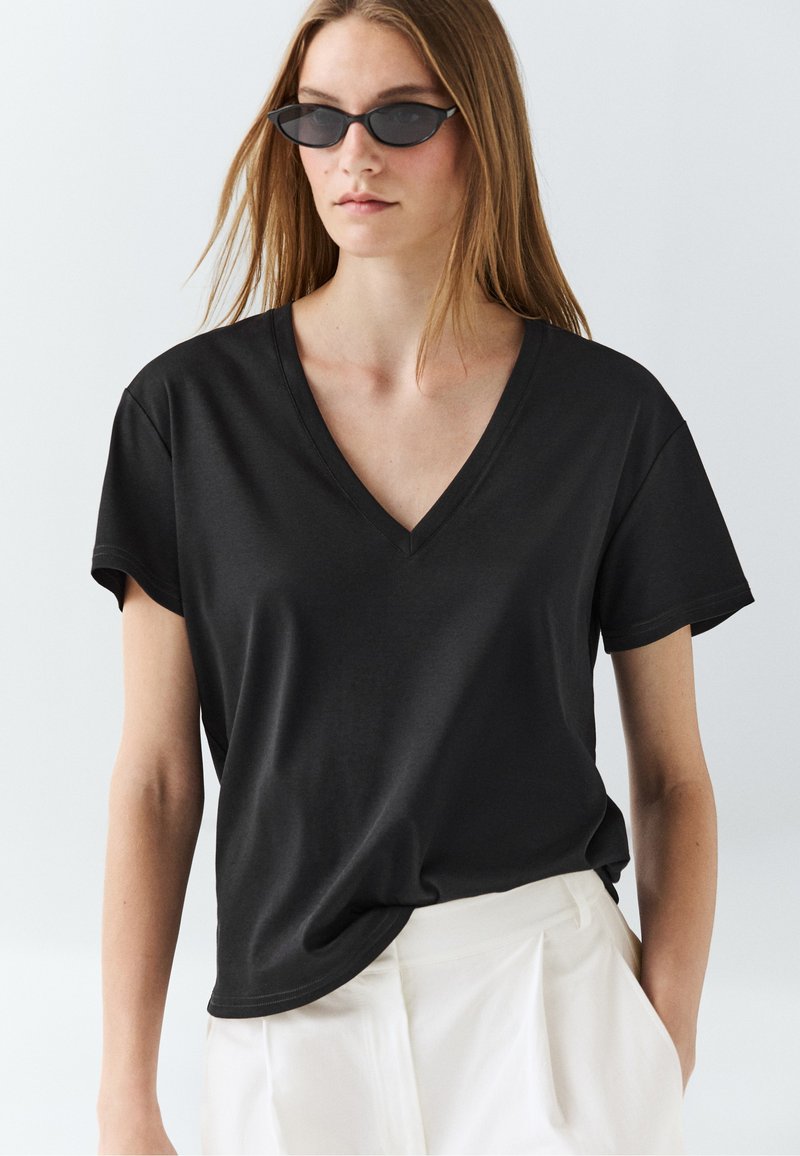 Junge Frau trägt ein schwarzes T-Shirt mit V-Ausschnitt, weiße hoch taillierte Hose und schmale schwarze Sonnenbrille, steht vor einem schlichten hellen Hintergrund.