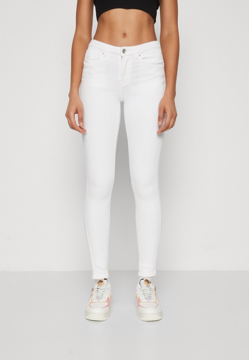 Femme portant un jean skinny blanc taille haute et des baskets blanches avec des accents roses, debout devant un fond clair uni.