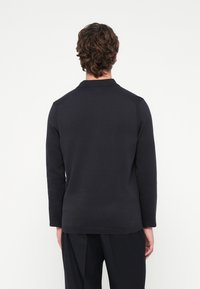 Tumši zils garām piedurknēm polos krekls ar gludu tekstūru, ar apkakli un plecu šuvēm, valkāts virs melnām biksēm.