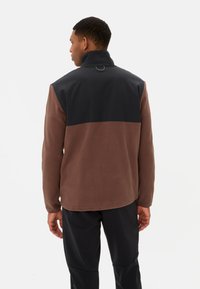 Giacca in pile marrone e nera con colletto alto, caratterizzata da un design bicolore e un accento circolare sul retro, con texture liscia.
